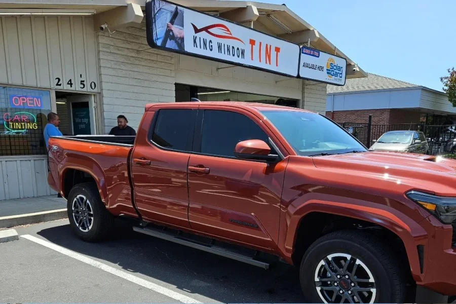 Orange 2024 Toyota Tacoma with freshly tinted windows, beside the King Window Tint sign on Fulton Ave, Sacramento